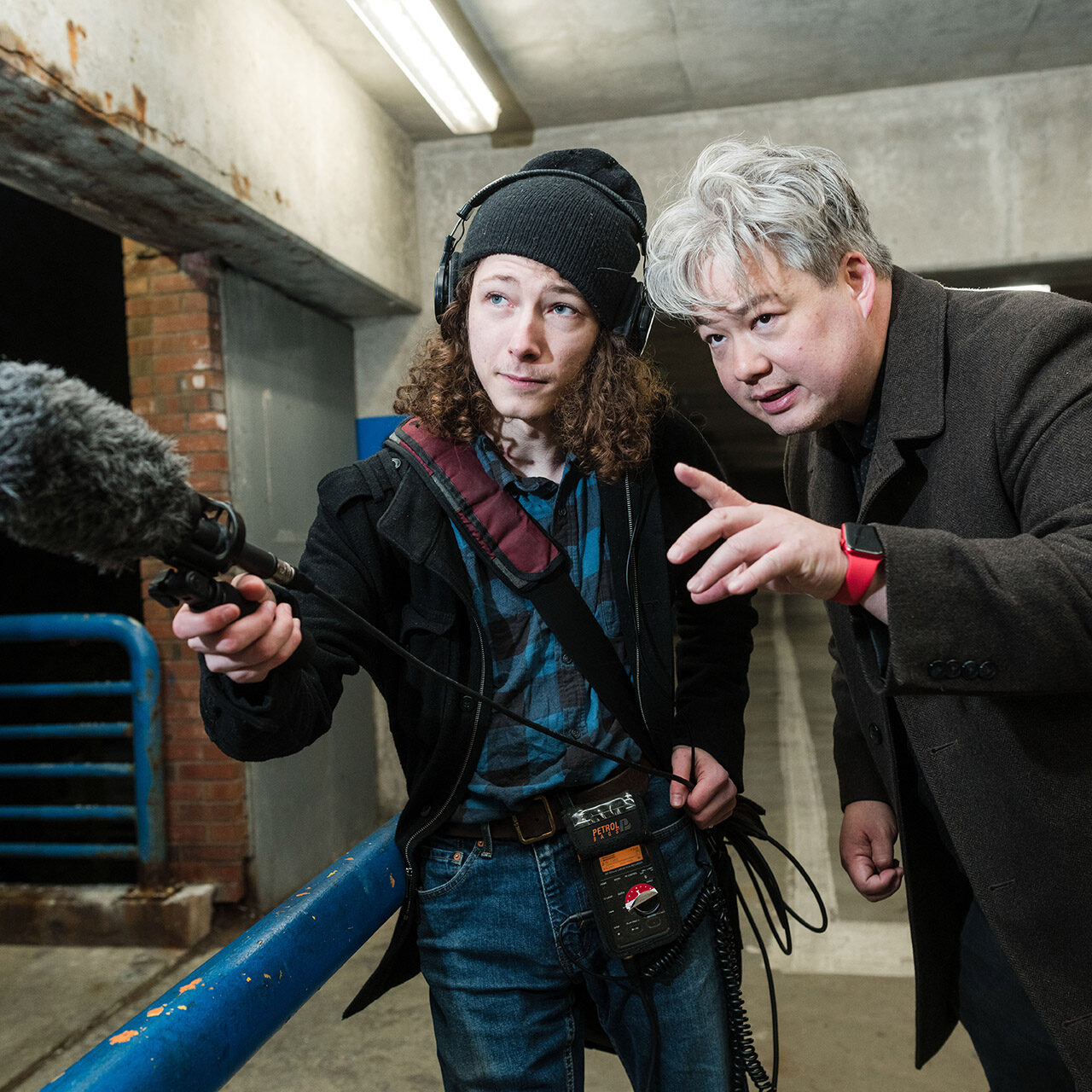 male student holds recording equipment in parking garage; male professor instructs